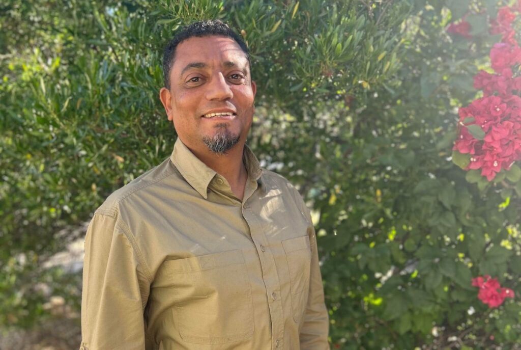 Romondo Jordani - The Brief | Namibia's Leading Business & Financial News Man in a khaki button-down shirt stands outdoors, smiling, with lush green foliage and pink flowers in the background.