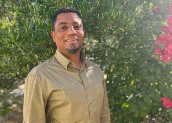 Man in a khaki button-down shirt stands outdoors, smiling, with lush green foliage and pink flowers in the background.