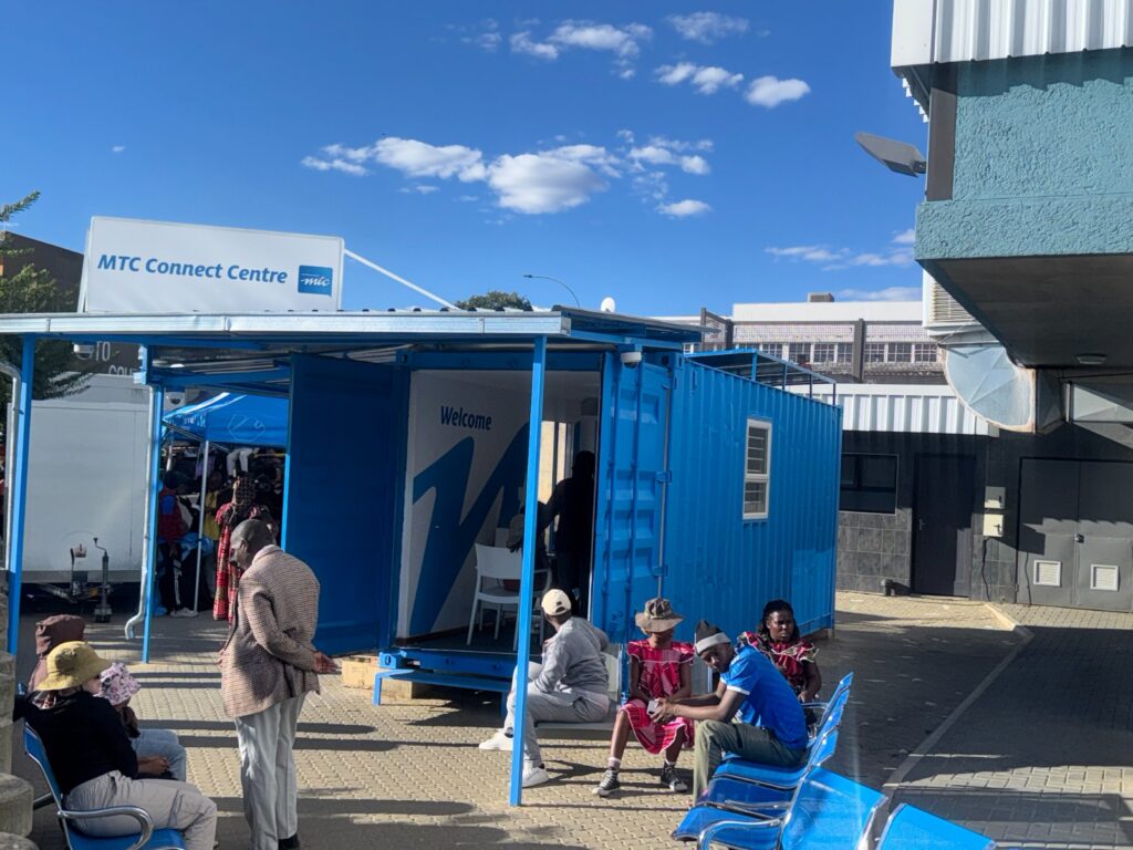 WhatsApp Image 2026-05-18 at 80009 AM - The Brief | Namibia's Leading Business & Financial News Outdoor community event beside a blue shipping-container booth marked 'Welcome' with attendees seated on blue chairs under a bright blue sky. (MTC Connect Centre sign visible)