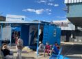 WhatsApp Image 2026-05-18 at 80009 AM - The Brief | Namibia's Leading Business & Financial News Outdoor community event beside a blue shipping-container booth marked 'Welcome' with attendees seated on blue chairs under a bright blue sky. (MTC Connect Centre sign visible)
