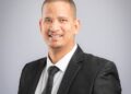 Smiling man in a dark suit and tie posing for a professional headshot against a gray backdrop.