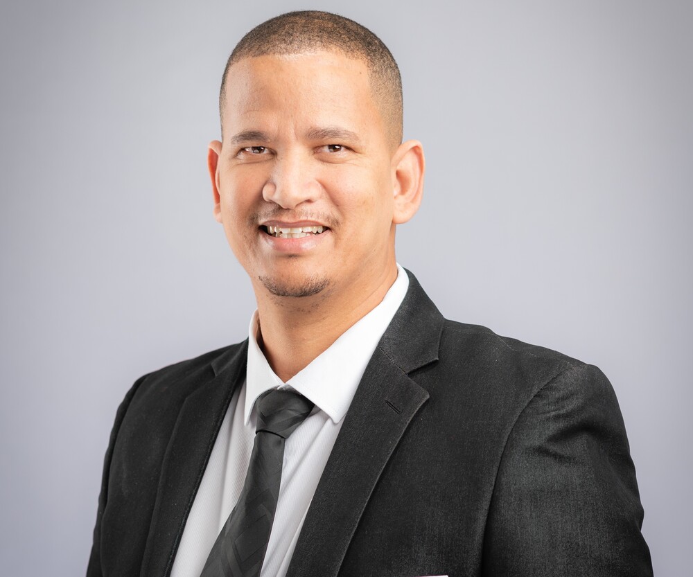 Smiling man in a dark suit and tie posing for a professional headshot against a gray backdrop.