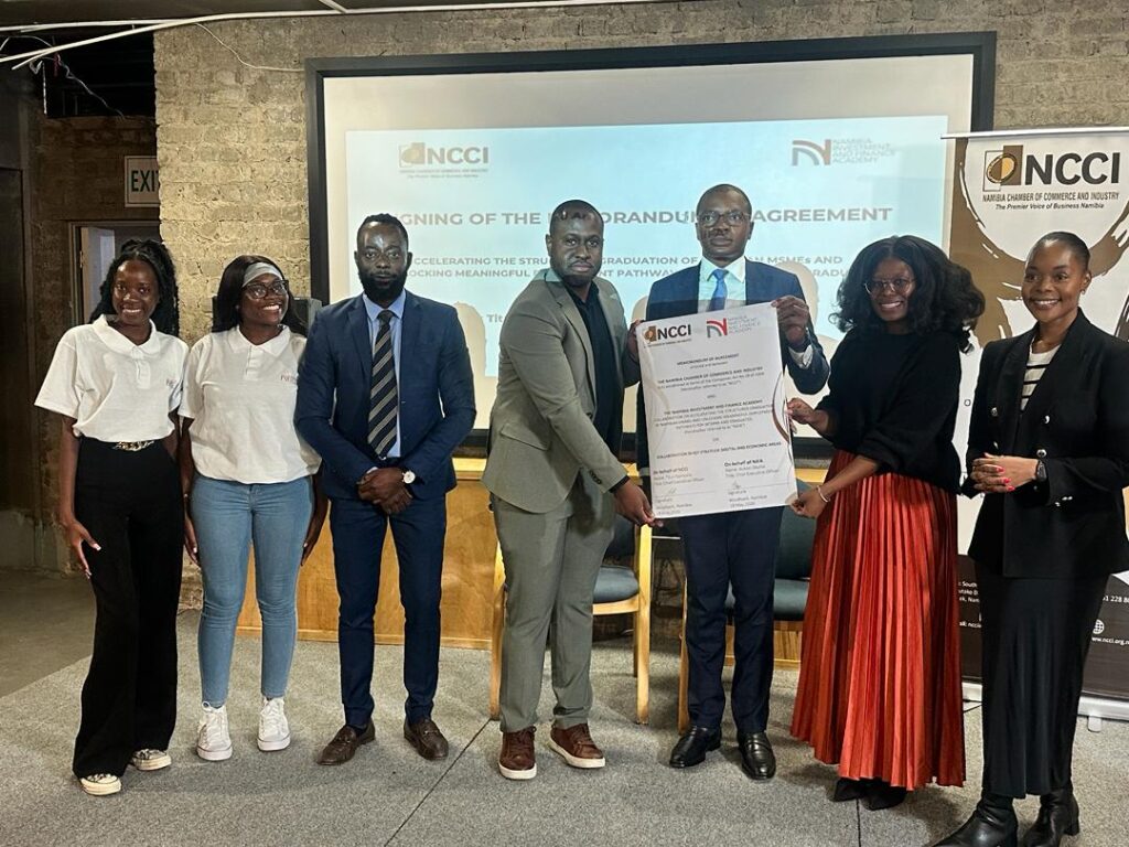 WhatsApp Image 2026-05-18 at 123924 PM - The Brief | Namibia's Leading Business & Financial News Group of seven professionals posing with a large document during a signing ceremony, in front of a projection screen and NCCI banners.