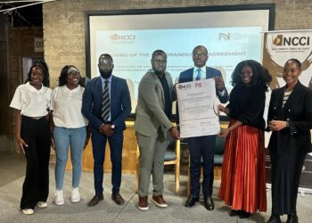 Group of seven professionals posing with a large document during a signing ceremony, in front of a projection screen and NCCI banners.