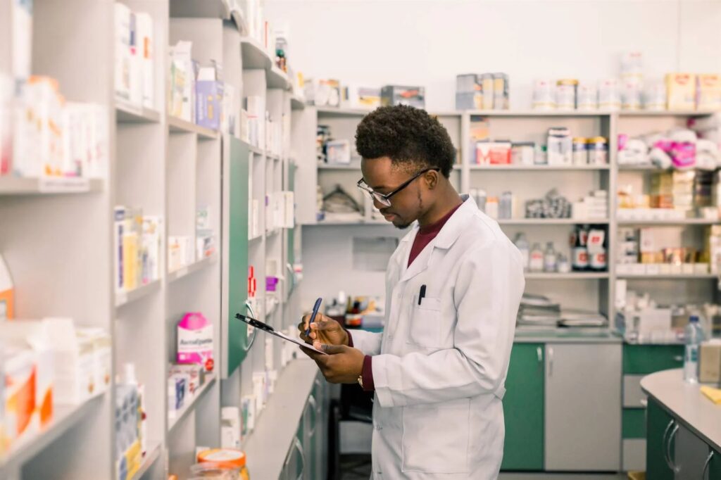 PHARMACIST - The Brief | Namibia's Leading Business & Financial News Pharmacist in a white coat writes on a clipboard in a well-stocked pharmacy aisle.