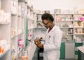 Pharmacist in a white coat writes on a clipboard in a well-stocked pharmacy aisle.