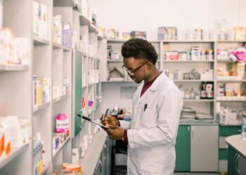 Pharmacist in a white coat writes on a clipboard in a well-stocked pharmacy aisle.