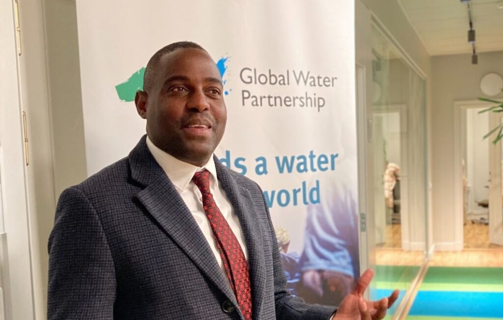 Alex Simalawi - The Brief | Namibia's Leading Business & Financial News Man in a dark suit and red tie standing beside a Global Water Partnership banner indoors.