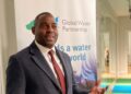 Alex Simalawi - The Brief | Namibia's Leading Business & Financial News Man in a dark suit and red tie standing beside a Global Water Partnership banner indoors.