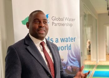 Man in a dark suit and red tie standing beside a Global Water Partnership banner indoors.
