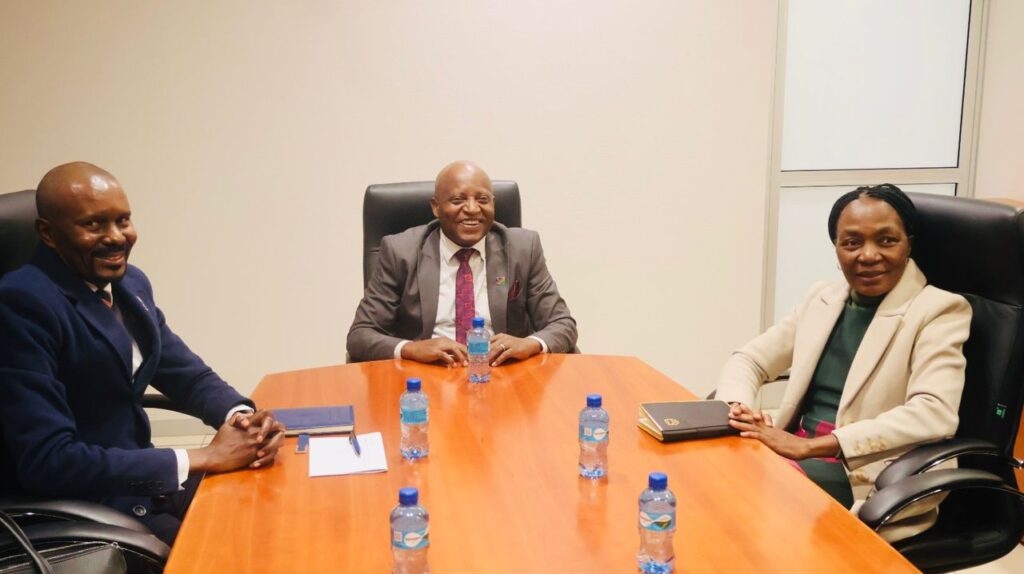Three professionals sit around a wooden conference table with water bottles and notebooks, smiling during a meeting in an office.