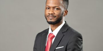Portrait of a man in a dark suit and red tie, looking at the camera against a plain gray background, with a white pocket square.