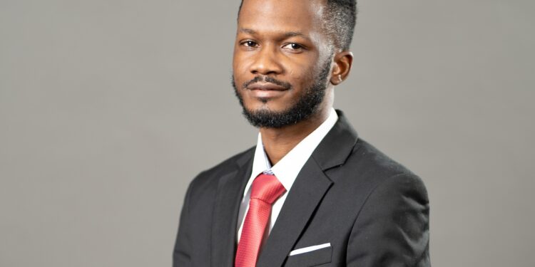 Portrait of a man in a dark suit and red tie, looking at the camera against a plain gray background, with a white pocket square.