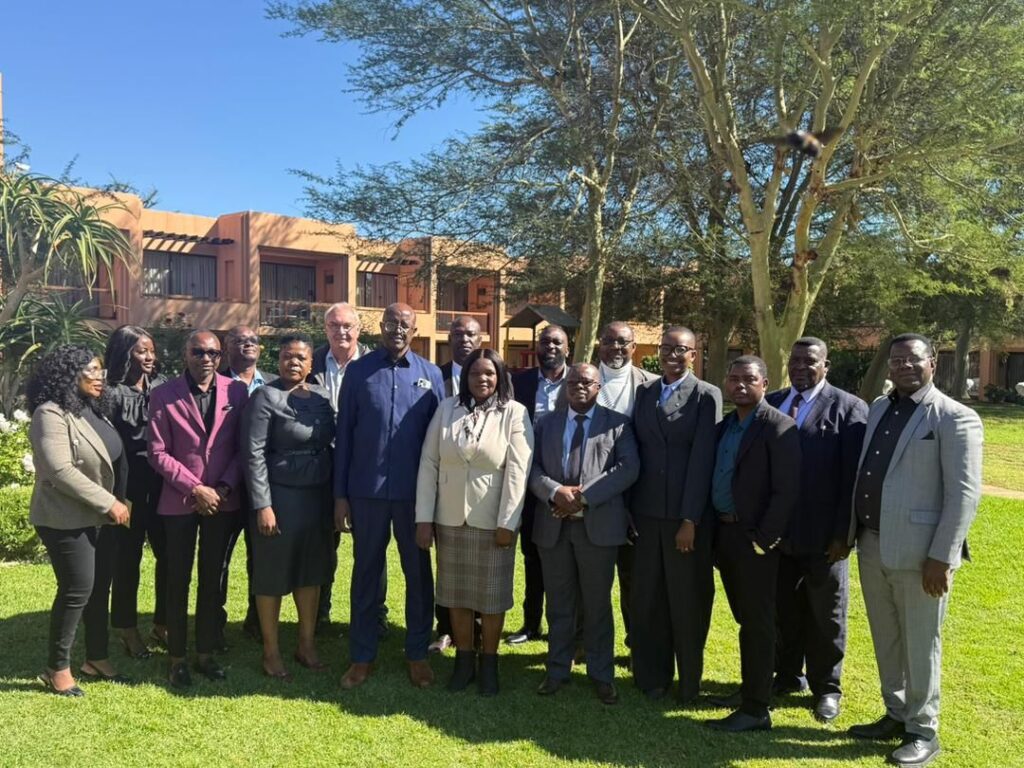 Group of professionals in business attire posing together on a sunny lawn with trees and a brick building in the background.