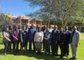 Group of professionals in business attire posing together on a sunny lawn with trees and a brick building in the background.