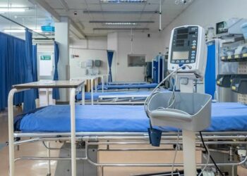 Hospital ward with blue-sheeted beds, privacy curtains, and a patient monitor in the foreground.