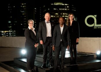 Four professionals in formal attire pose on a rooftop stage at night with city lights behind them.