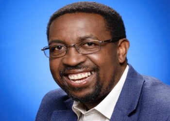Portrait of a smiling man wearing glasses and a blue blazer against a blue gradient background, looking at the camera.