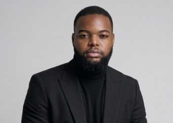 Professional head-and-shoulders portrait of a man in a black blazer and turtleneck against a gray background.