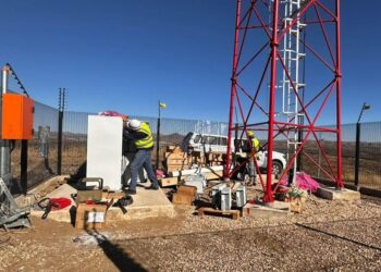 Two workers in hard hats and hi-vis vests install equipment next to a red lattice communications tower in a fenced, arid site.