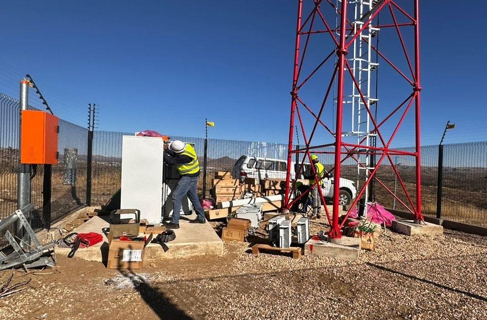 TOWER1 - The Brief | Namibia's Leading Business & Financial News Two workers in hard hats and hi-vis vests install equipment next to a red lattice communications tower in a fenced, arid site.