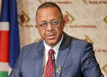 Man in a blue suit and red patterned tie speaks into a microphone at a podium, with a Namibian flag and Bank of Namibia backdrop behind him.
