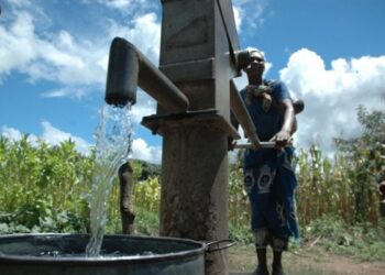 Man standing beside a rusted hand water pump, water pouring into a metal barrel in a rural field under a bright blue sky.