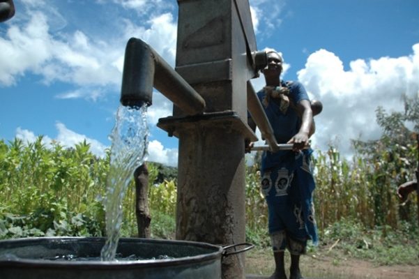 water no2 - The Brief | Namibia's Leading Business & Financial News Man standing beside a rusted hand water pump, water pouring into a metal barrel in a rural field under a bright blue sky.
