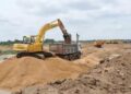 Yellow excavator loading dirt into a dump truck at a construction site, with a sandy trench and distant equipment on the horizon.