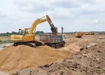 Yellow excavator loading dirt into a dump truck at a construction site, with a sandy trench and distant equipment on the horizon.