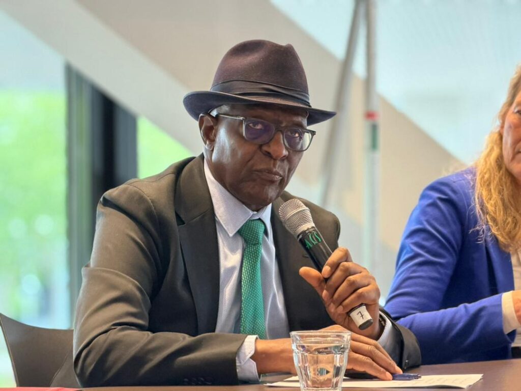 Man in a dark suit and green tie wearing a hat, speaking into a handheld microphone at a panel discussion