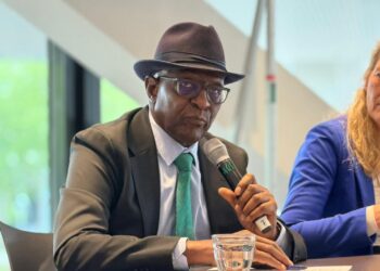 Man in a dark suit and green tie wearing a hat, speaking into a handheld microphone at a panel discussion
