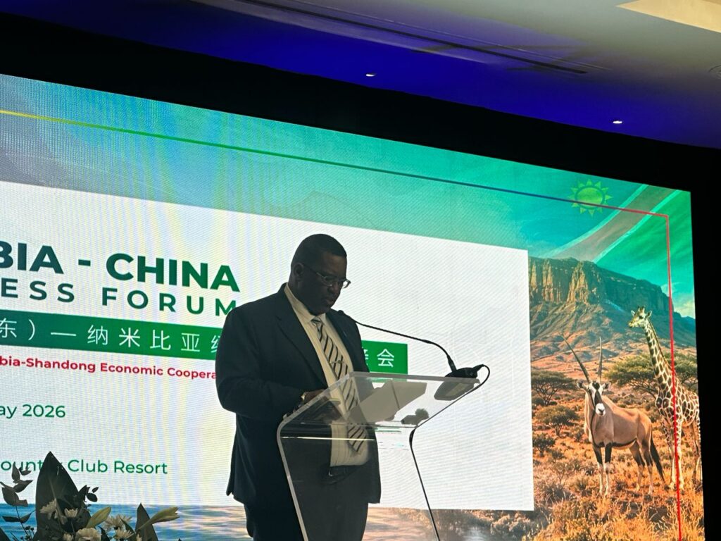 Charles Josob - The Brief | Namibia's Leading Business & Financial News Man in a suit speaks at a clear podium during a Zambia–China Business Forum, with a large screen backdrop showing landscape and wildlife mascots behind him.