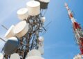 Tall communication tower with numerous white satellite dishes and antennas against a clear blue sky in the foreground.