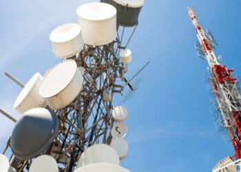 Tall communication tower with numerous white satellite dishes and antennas against a clear blue sky in the foreground.