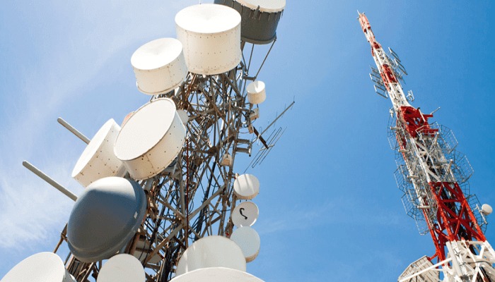 Tall communication tower with numerous white satellite dishes and antennas against a clear blue sky in the foreground.