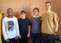 Four men stand side by side, smiling for a casual group photo in an office-like room with a brown wall behind them.