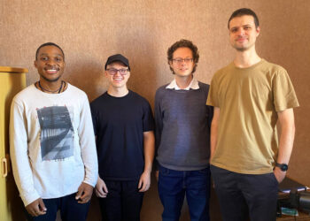 Four men stand side by side, smiling for a casual group photo in an office-like room with a brown wall behind them.