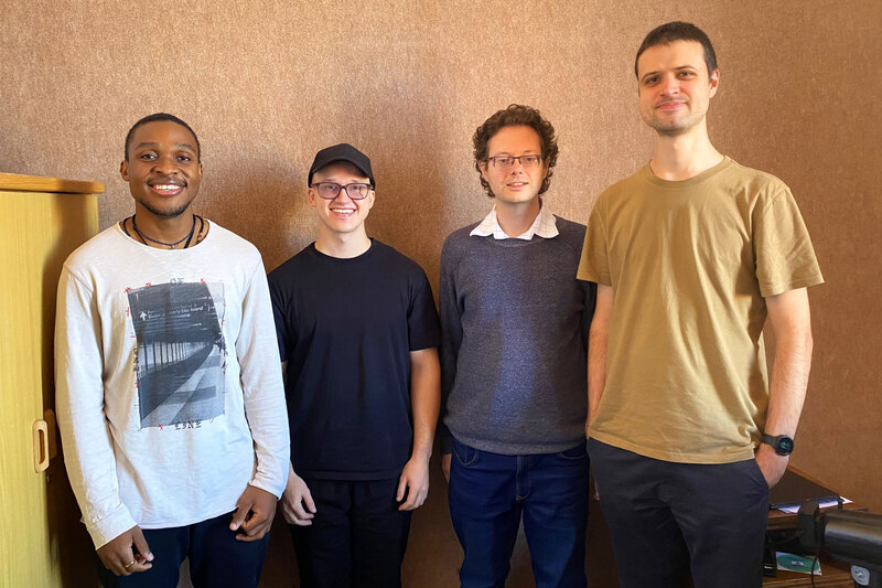 Four men stand side by side, smiling for a casual group photo in an office-like room with a brown wall behind them.