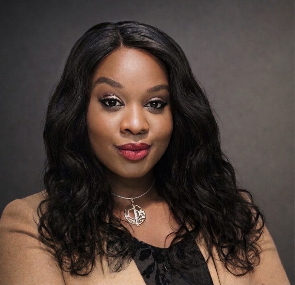 Portrait of a woman with long wavy black hair, wearing a beige blazer and a silver pendant necklace, smiling softly against a dark gray backdrop.