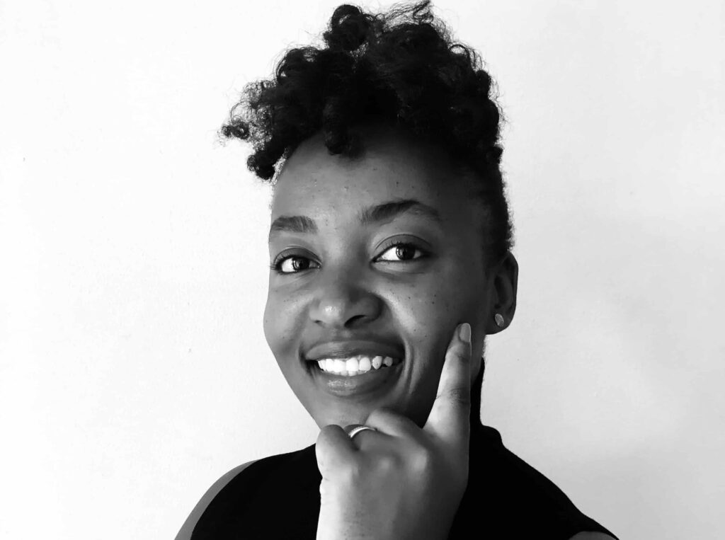 Asteria Pirola - The Brief | Namibia's Leading Business & Financial News Close-up black-and-white portrait of a smiling woman with a curly top knot, hand resting on her chin.