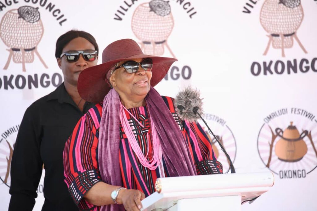 WhatsApp Image 2026-05-27 at 21035 PM - The Brief | Namibia's Leading Business & Financial News Elderly woman in a wide-brimmed hat and sunglasses speaks at a podium during a festival, with a security guard in black behind her.
