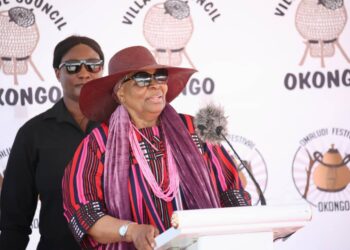 Elderly woman in a wide-brimmed hat and sunglasses speaks at a podium during a festival, with a security guard in black behind her.