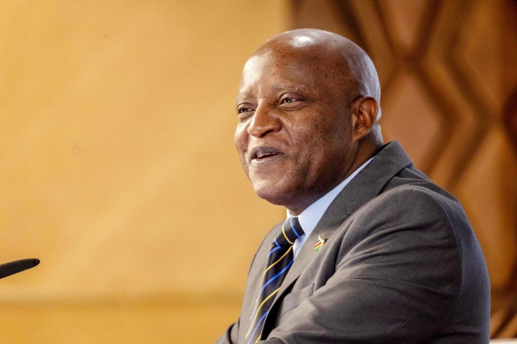 PRIME MINISTER - The Brief | Namibia's Leading Business & Financial News Man in a dark suit and striped tie speaks at a podium; a microphone is visible nearby.