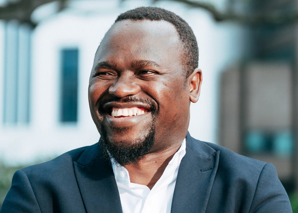 George SANZILA - The Brief | Namibia's Leading Business & Financial News Close-up portrait of a smiling Black man in a dark suit outdoors, white shirt visible beneath.