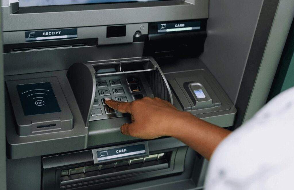 Hand entering PIN on an ATM keypad with card slot and cash dispenser visible.
