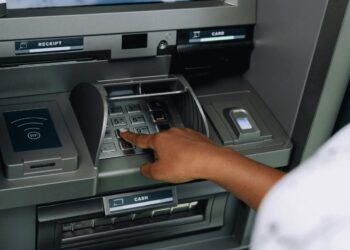 Hand entering PIN on an ATM keypad with card slot and cash dispenser visible.
