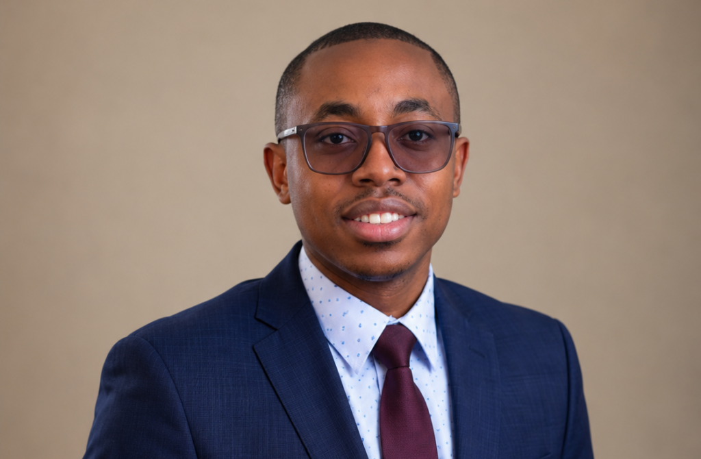 Professional headshot of a man in a navy suit, white dotted shirt, burgundy tie, and glasses, smiling at the camera.