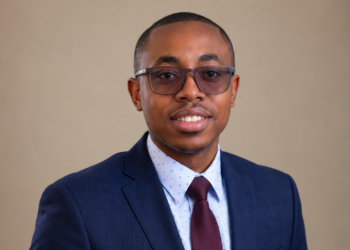 Professional headshot of a man in a navy suit, white dotted shirt, burgundy tie, and glasses, smiling at the camera.