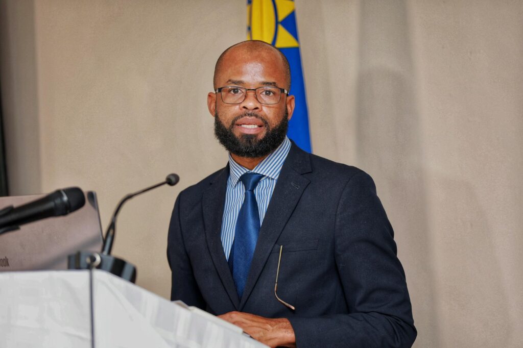 Stanley Shanapinda1 - The Brief | Namibia's Leading Business & Financial News Man in a navy suit and blue striped shirt speaks at a podium with a microphone, flag in background.